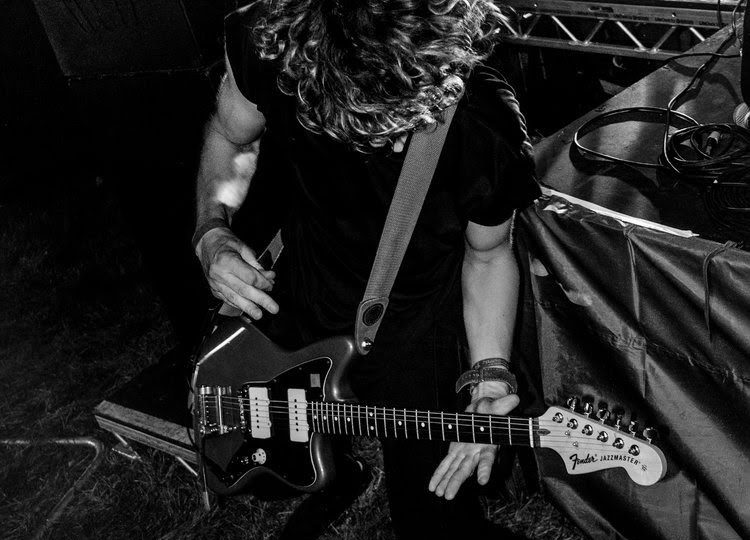 A white, masculine person with medium length curly hair and wearing a black top playing an electric guitar.