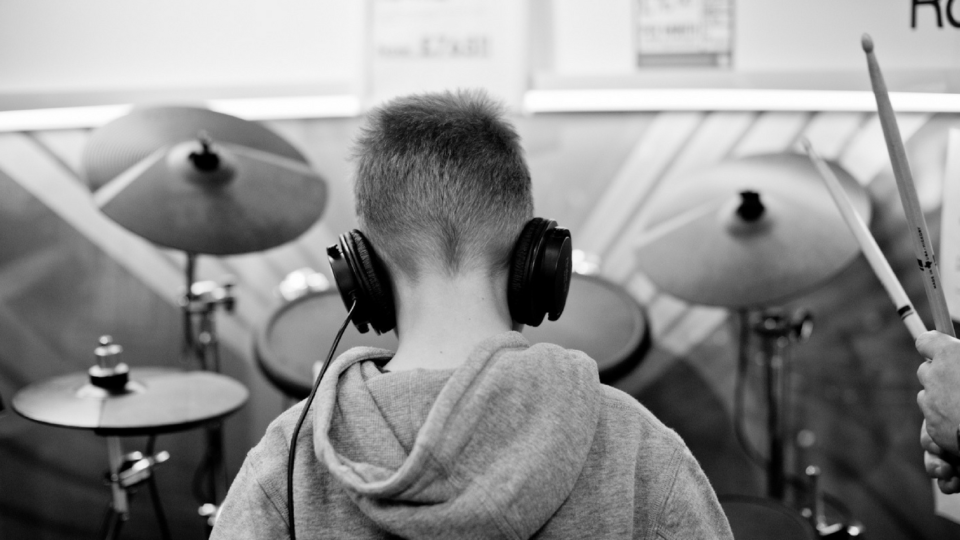 A white, masculine person with short hair and wearing a grey jumper playing a drum kit with headphones on.