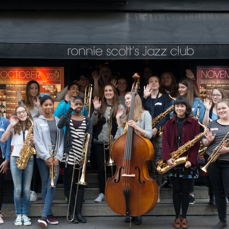 A group of people with mixed identities standing outside "Ronnie Scott's Jazz Club", all holding a jazz instrument.