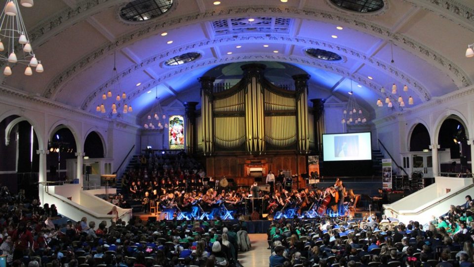 A group of people with mixed identities watching an orchestra of mixed identities and a playing range of instruments.