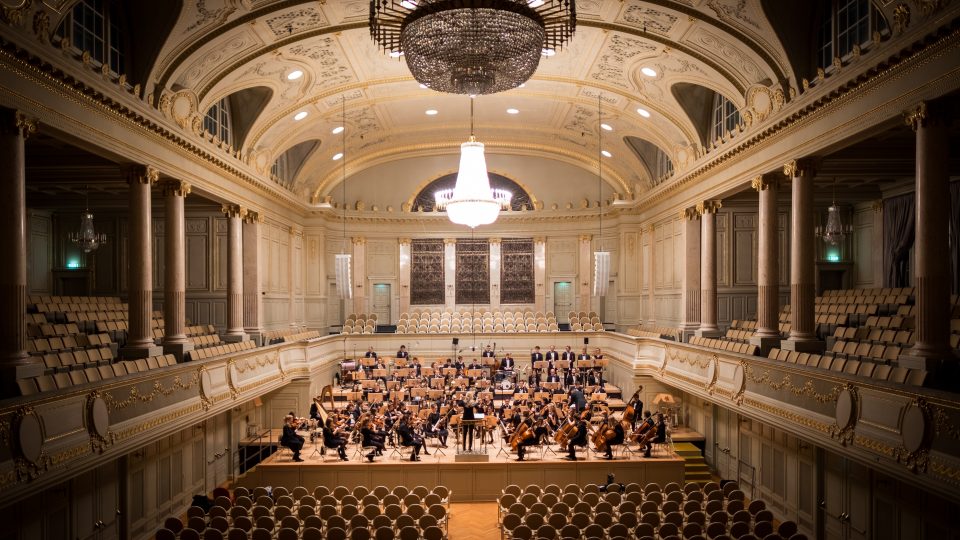 An orchestra playing a range of instruments in an orchestra hall.