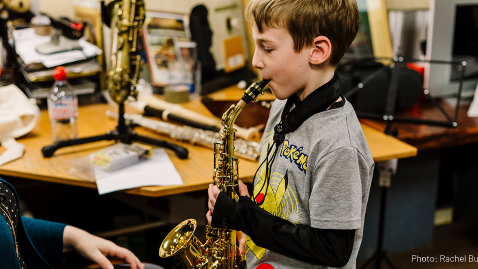 A young, white, masculine child with short, brown hair and wearing a grey Pokémon top who is playing a saxophone