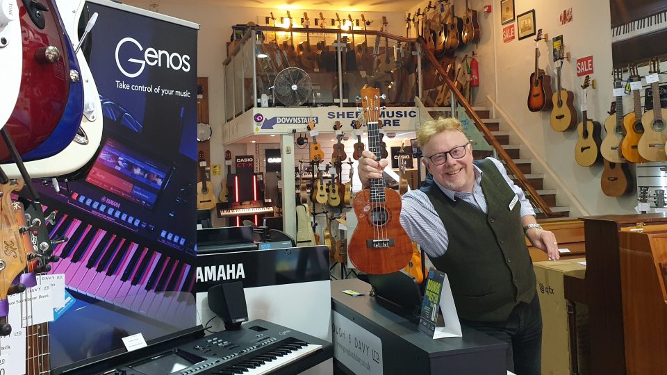 A white, masculine adult with an excited expression, yellow hair and wearing a waistcoat is waving a ukulele in a shop