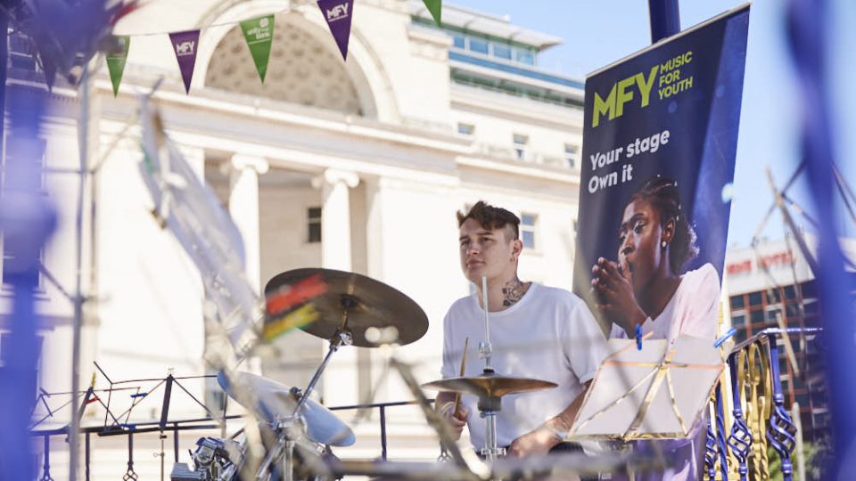 A white, masculine person with short, brown hair and wearing a white top is playing the drums