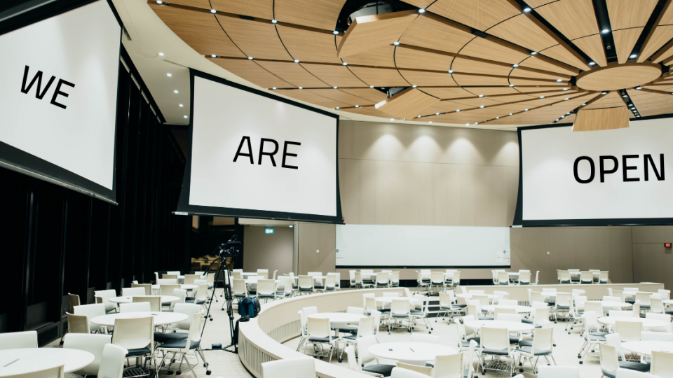 A room with lots of tables an chairs and some projector screens that say "WE ARE OPEN"