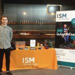 A white, masculine adult with short, brown hair and wearing a checkered shirt is standing in front of an "ISM" stand