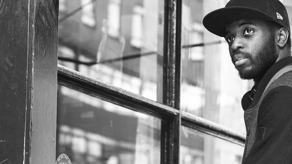 A black, masculine adult wearing a baseball cap and a black and grey jumper is standing outside a shop.