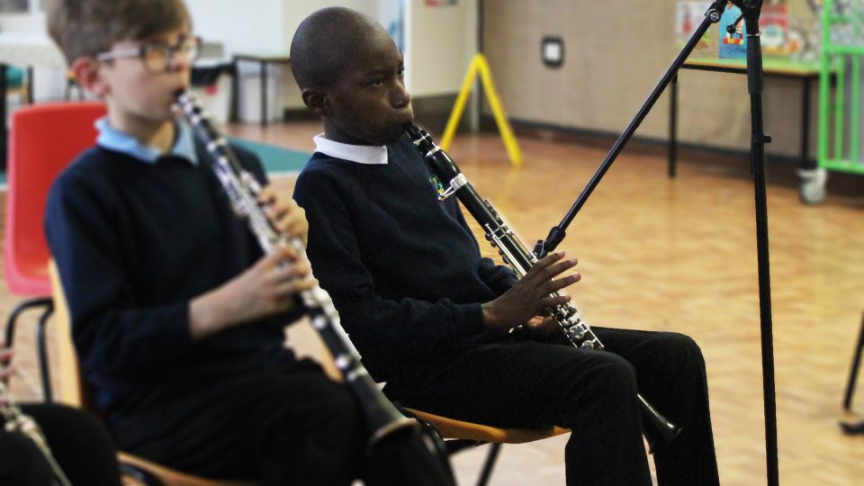 A bald, black, masculine child who is wearing a blue school jumper and black trousers is playing a clarinet with one hand.