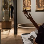 A brown, feminine adult sitting with a guitar in an art gallery and studying a piece of music