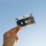 A white person holding a cassette tape with a blue sky background