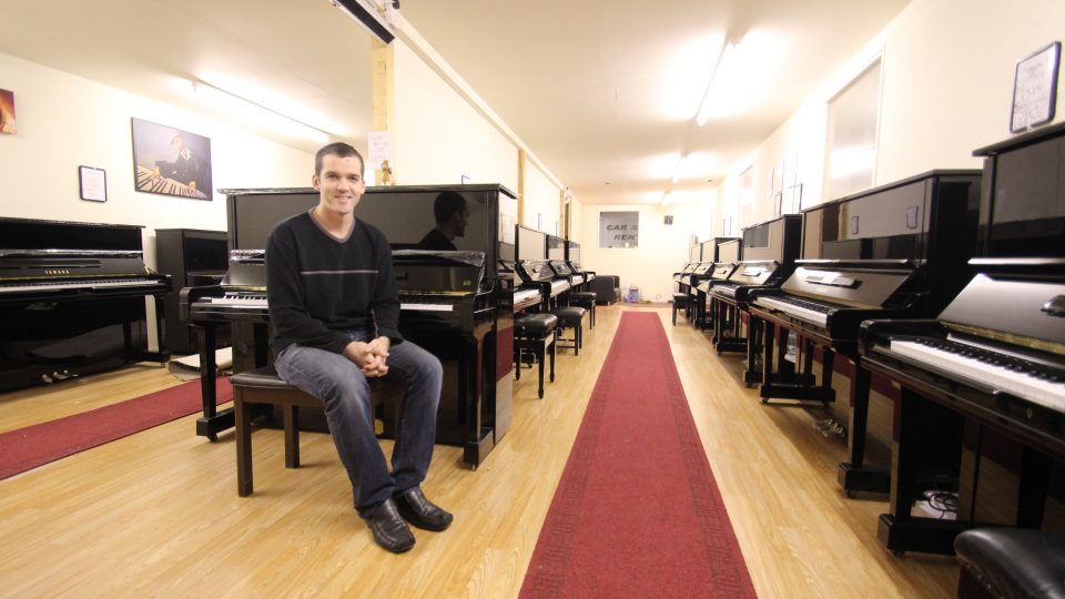 A white, masculine adult with short, brown hair and wearing a black top is sitting on a stool in a room of upright pianos.