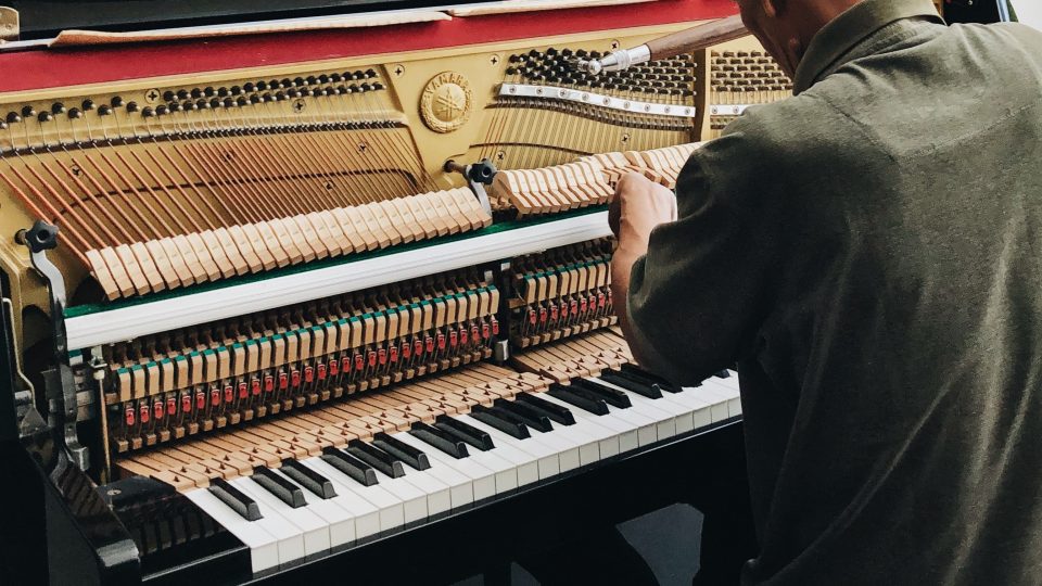 An elderly, white, masculine adult tuning a piano