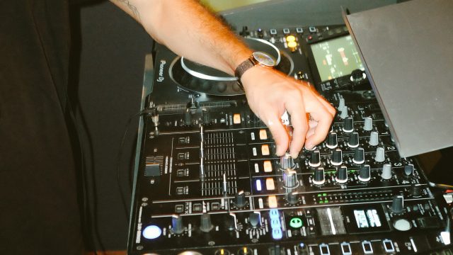 A white, masculine adult with a black top and beige shorts is using a DJ turntable