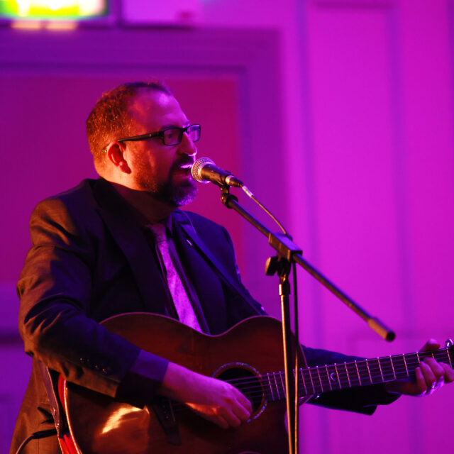 A white, masculine adult with short, brown hair and beard and wearing a suit is playing an acoustic guitar and singing